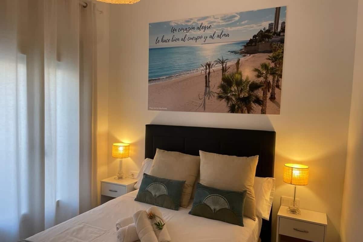 Een netjes opgemaakt bed met kussens en een handdoek, een nachtkastje met een lamp, en een muur met een strandlandschap en tekst.