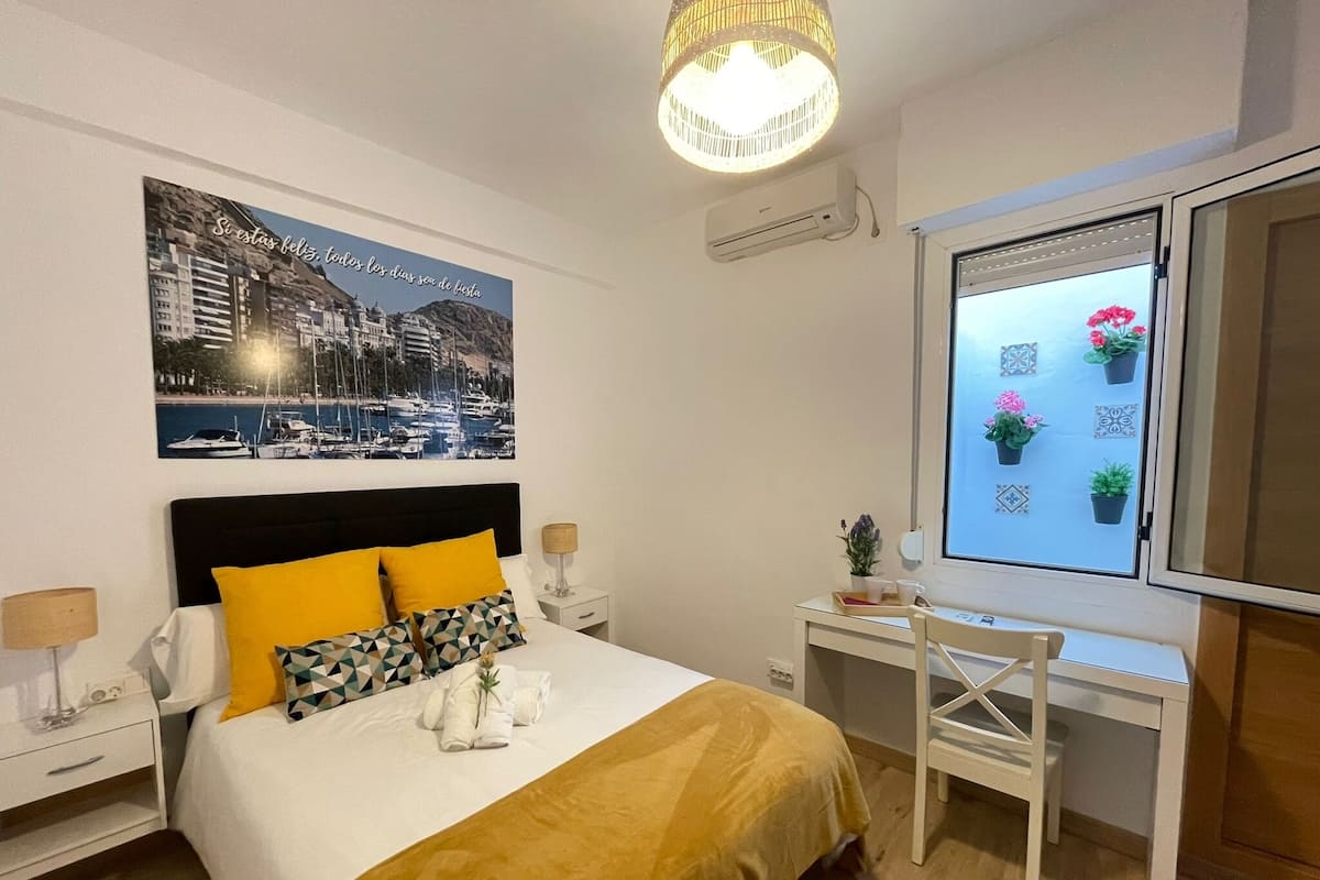Hotelkamer met een bed, een bureau, een stoel, een raam met plantenbakken en een wandairconditioning.