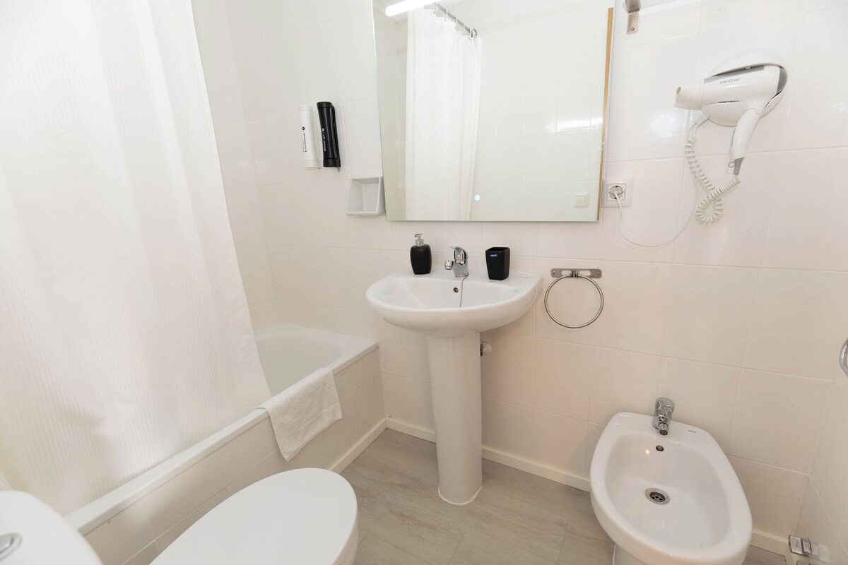 A modern bathroom with a bathtub, a pedestal sink, a bidet, and a hairdryer mounted on the wall.