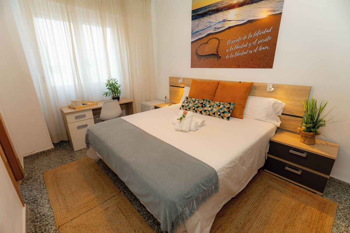 A neatly arranged bedroom with a bed, bedside tables, a desk, and a window with sheer curtains.