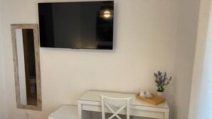 A room with a white desk, a small white chair, a television mounted on the wall, a small white refrigerator, and a mirror on the wall. - Hostal Ventura Premium  (Alicante)