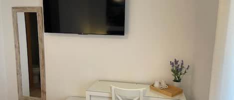 A room with a white desk, a small white chair, a television mounted on the wall, a small white refrigerator, and a mirror on the wall.