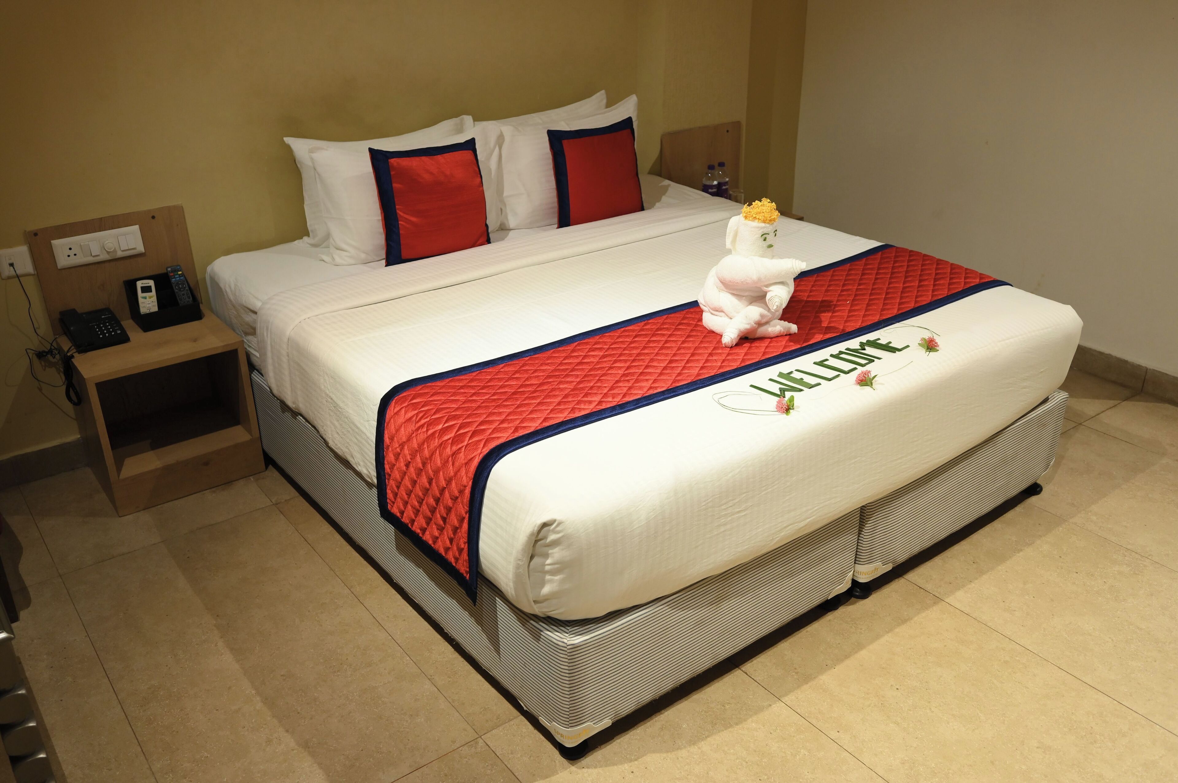 A neatly made bed with a red quilt, a plush white teddy bear, and a welcome mat.