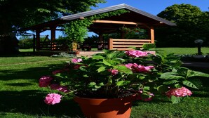 Gazebo amb teulada de fusta i una panxa, envoltat de vegetació exuberant i flors roses.