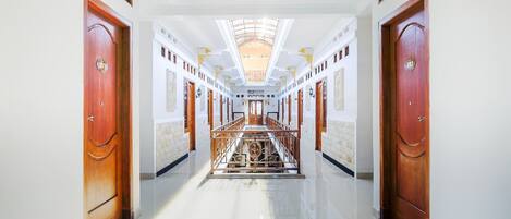 A long, well-lit hallway with a central staircase, flanked by wooden doors and white walls.
