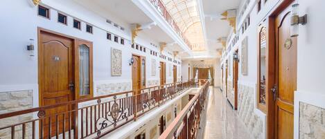 A grand hotel corridor with ornate railings, multiple doors, and a high ceiling with a skylight.
