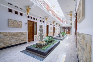 Un pasillo con pisos de mármol, paredes de piedra y puertas de madera. Se observan estatuas y plantas en macetas a lo largo del sendero.