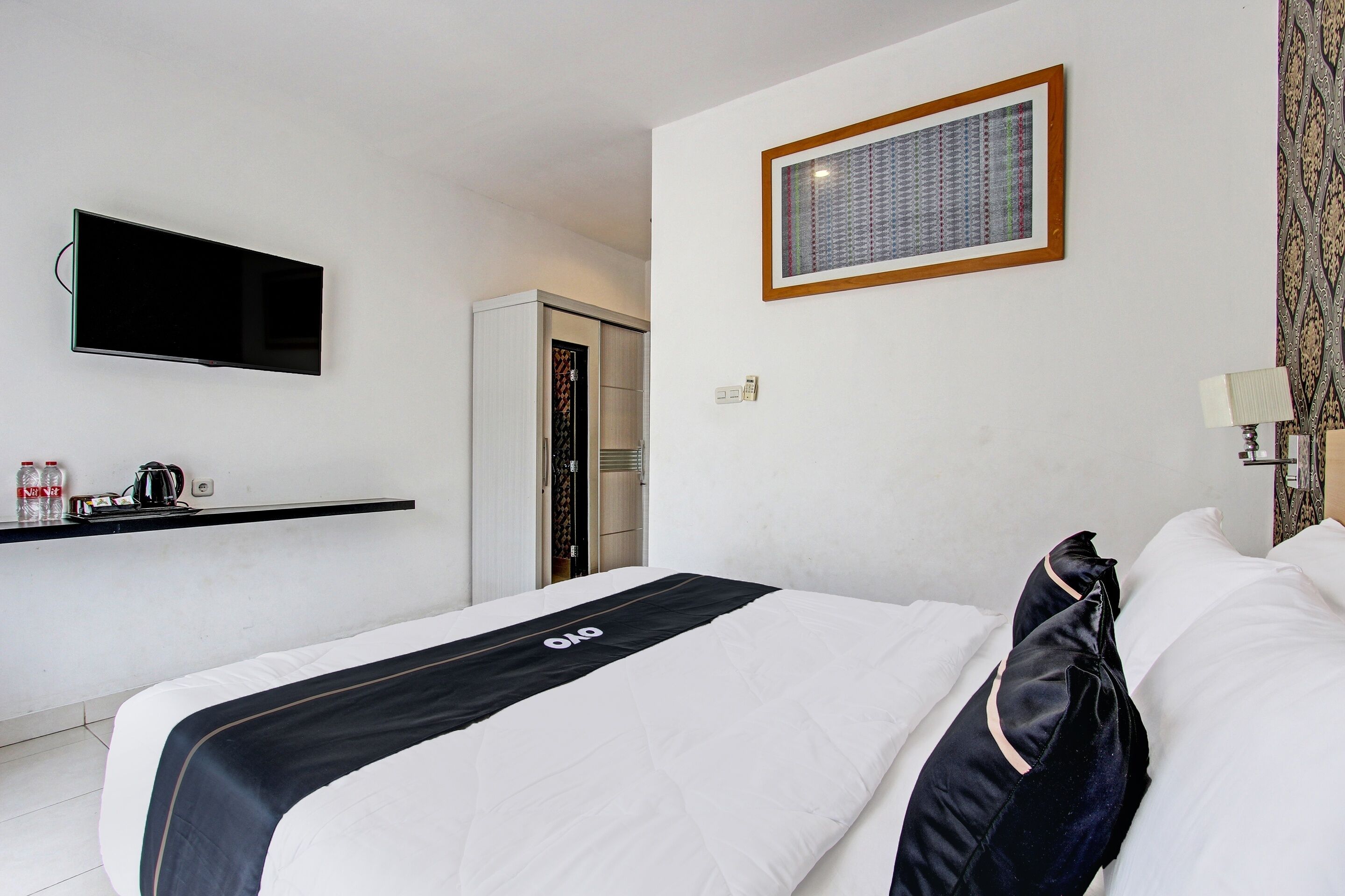 Habitación de hotel con una cama, un televisor de pantalla plana montado en la pared, una mesita con un hervidor de agua y un cuadro enmarcado.