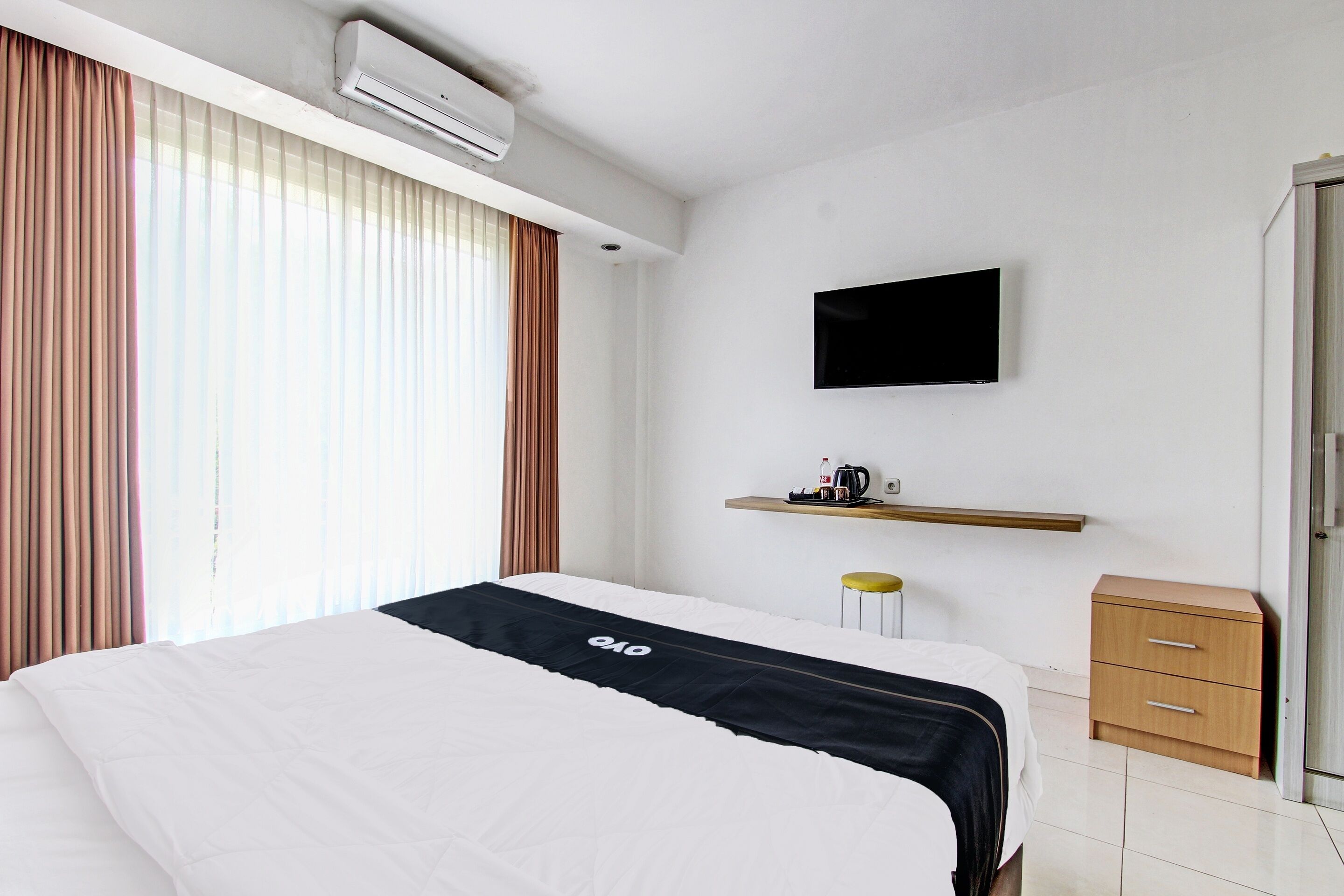 A hotel room with a bed, an air conditioner, a mounted TV, a small wooden nightstand, and a window with curtains.