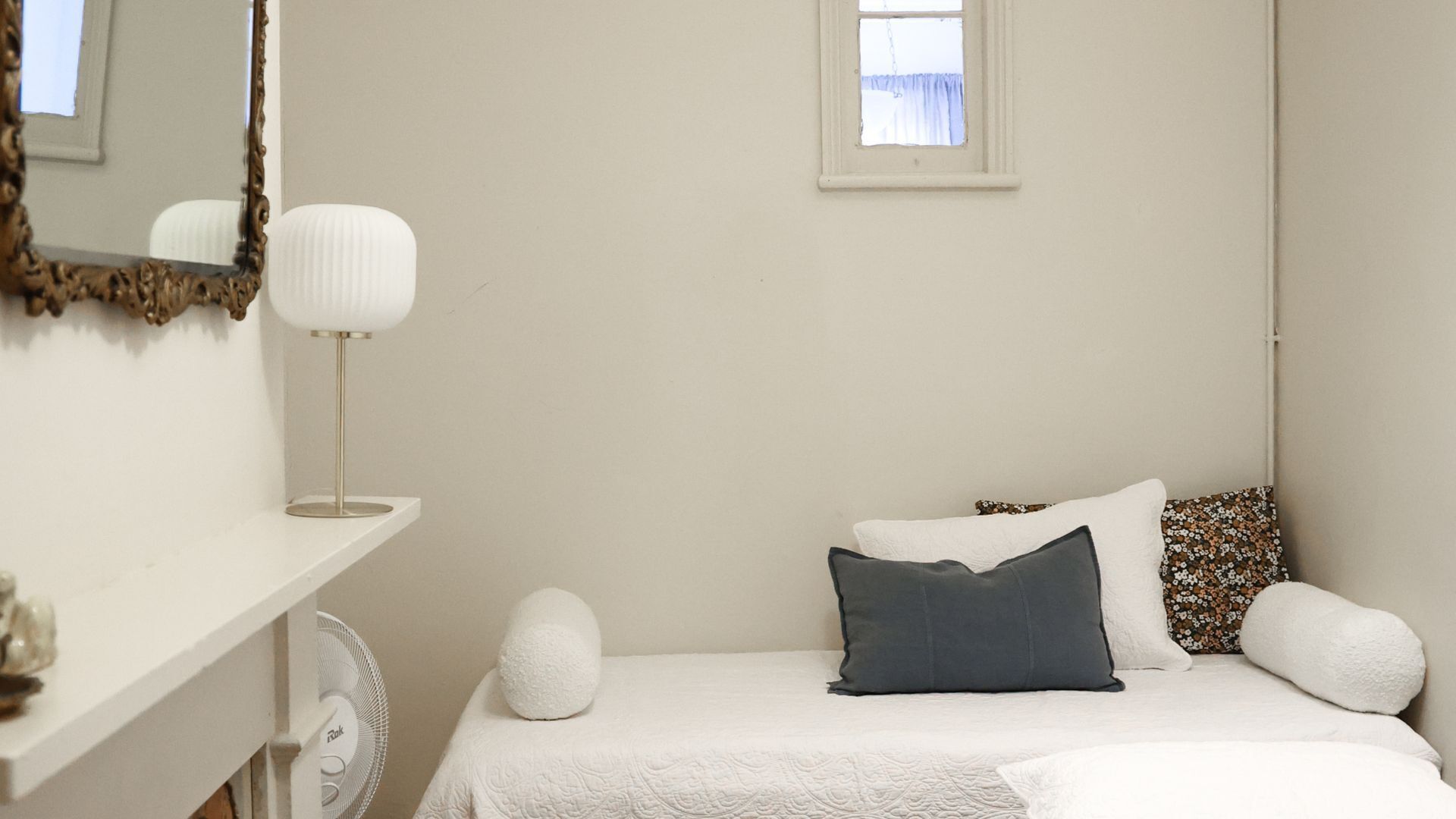 A bedroom with a bed, pillows, a bedside table, a lamp, a mirror, and a window.