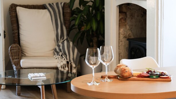 Una sala de estar acogedora con una mesa de centro de vidrio, un sofá de mimbre y una mesa redonda de madera con comida y copas de vino.