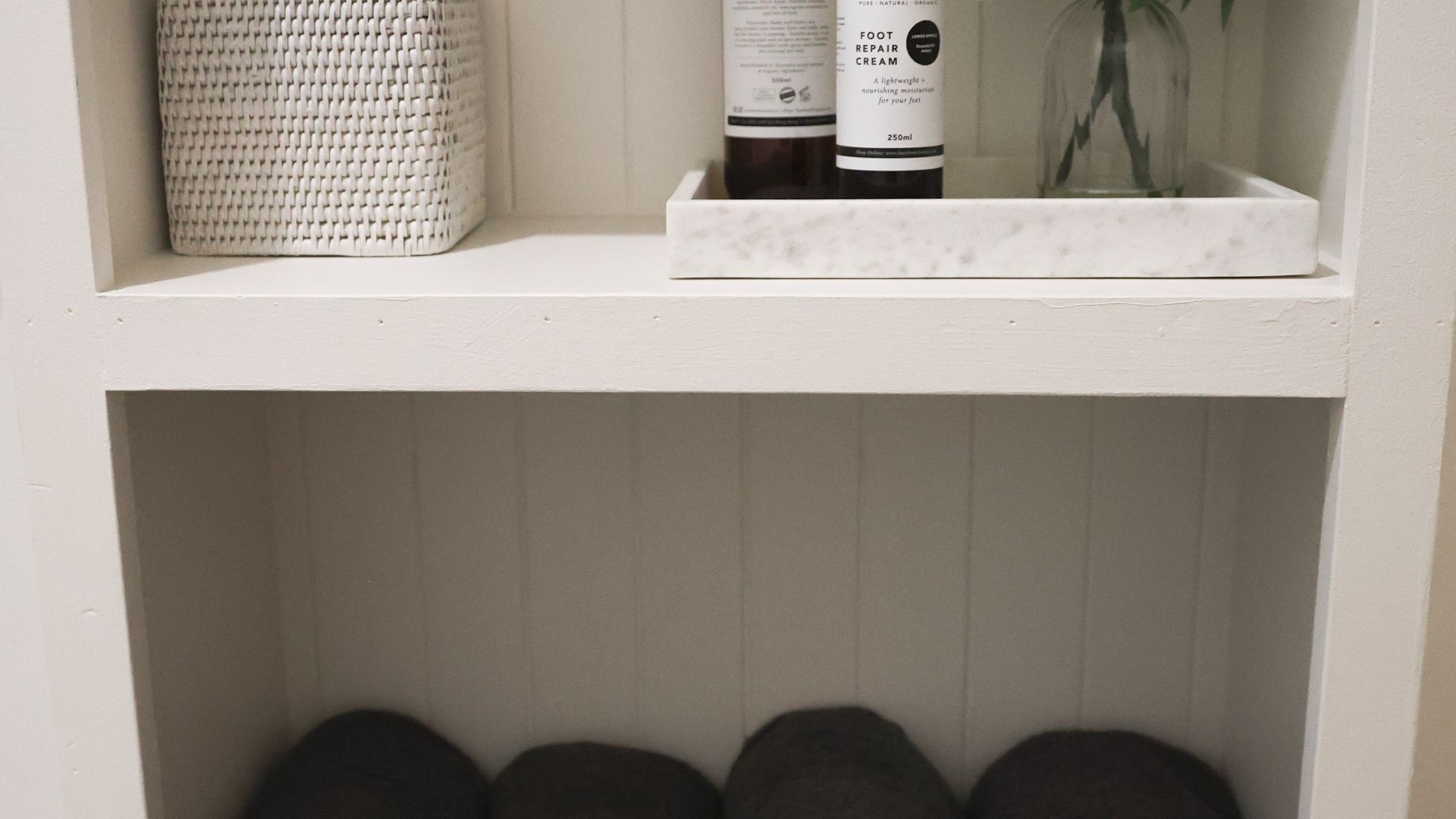 A shelf with a woven basket, two bottles of foot repair cream, and a glass container with a plant.