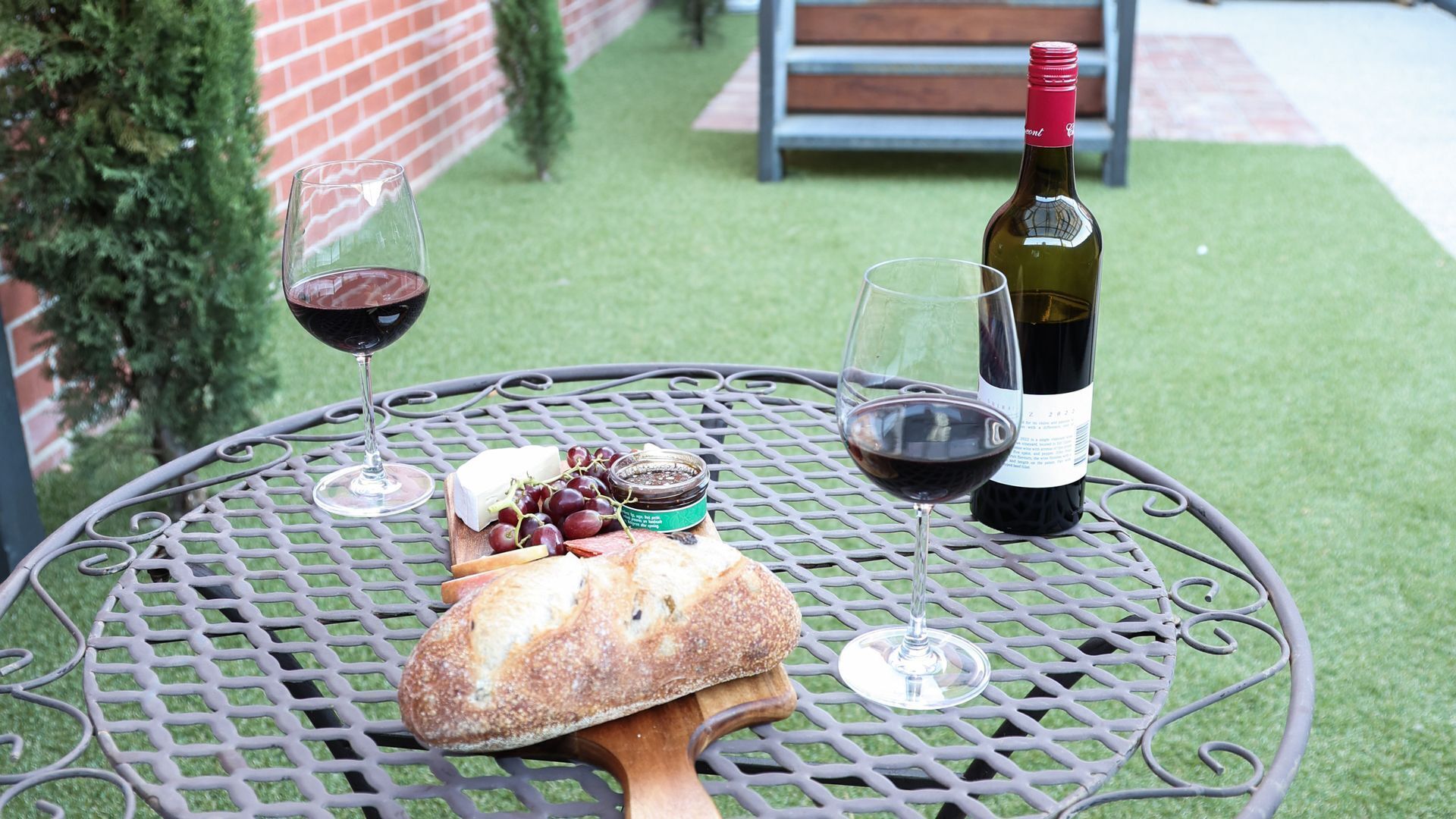 Una mesa redonda de metal con una botella de vino, dos copas de vino, una hogaza de pan y un frasco pequeño de mermelada.