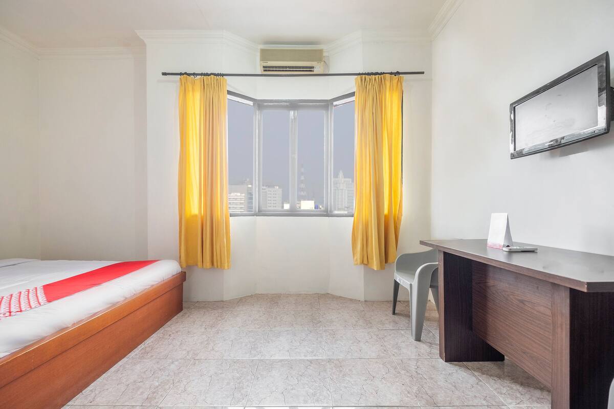 A hotel room with a bed, a desk, a chair, a window with curtains, and a mounted TV.