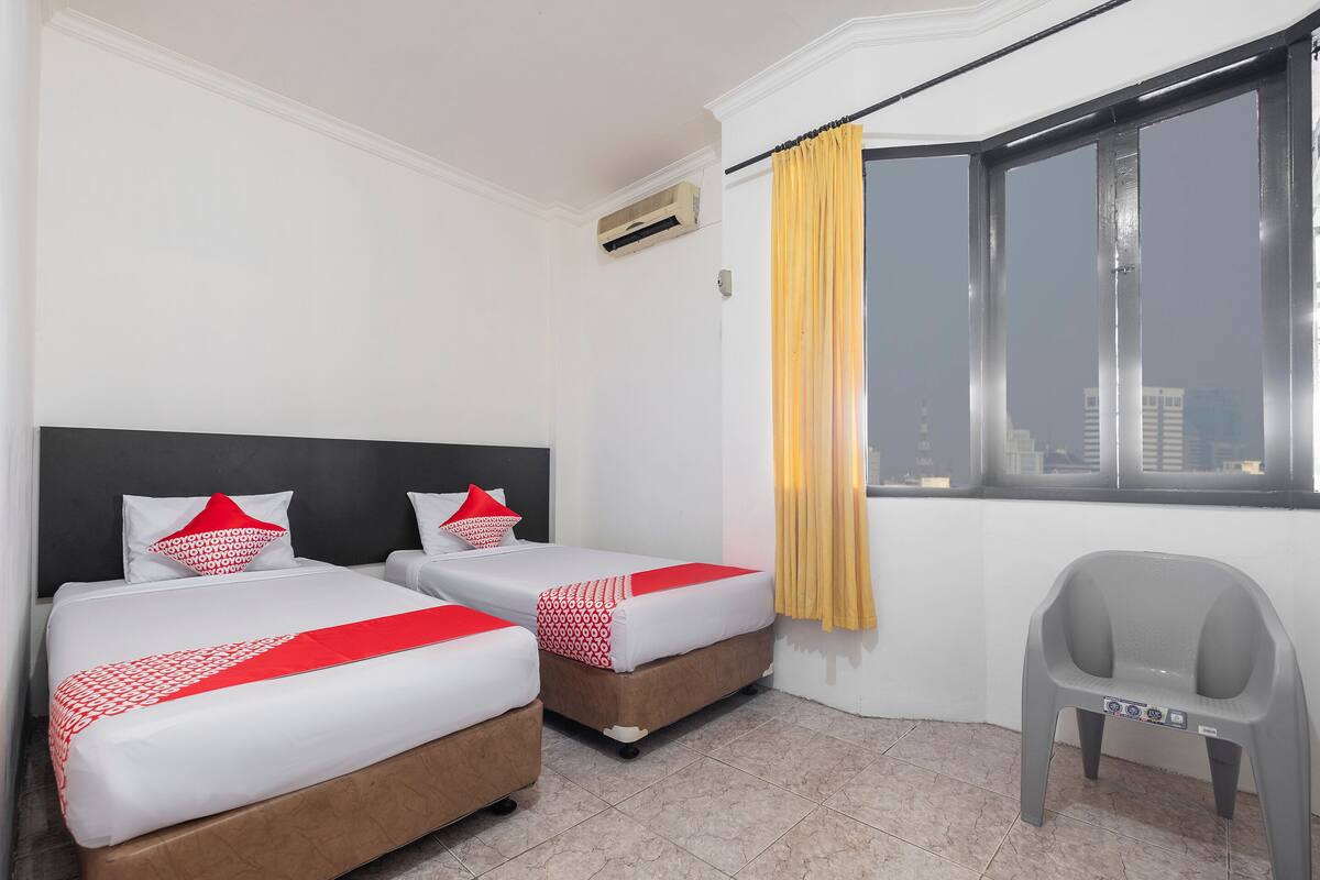 A hotel room with two beds, a window with yellow curtains, a gray plastic chair, and an air conditioning unit.