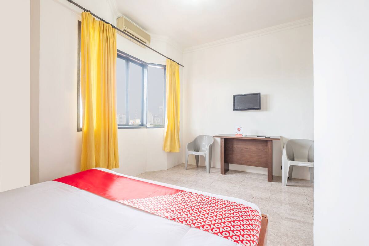 A hotel room with a bed, a desk, two chairs, a TV, and a window with curtains.