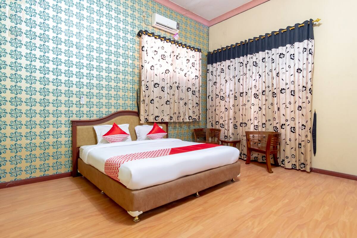 A neatly made bed with a red and white blanket, a wooden headboard, and two red pillows. There is a wooden chair and a small table in the corner. The room has patterned wallpaper and floral curtains.
