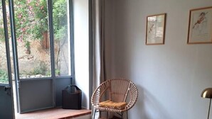 Une pièce dotée d’une grande porte-fenêtre coulissante donnant sur un jardin, d’une table basse en bois, d’un fauteuil en osier et de deux œuvres d’art encadrées accrochées au mur.