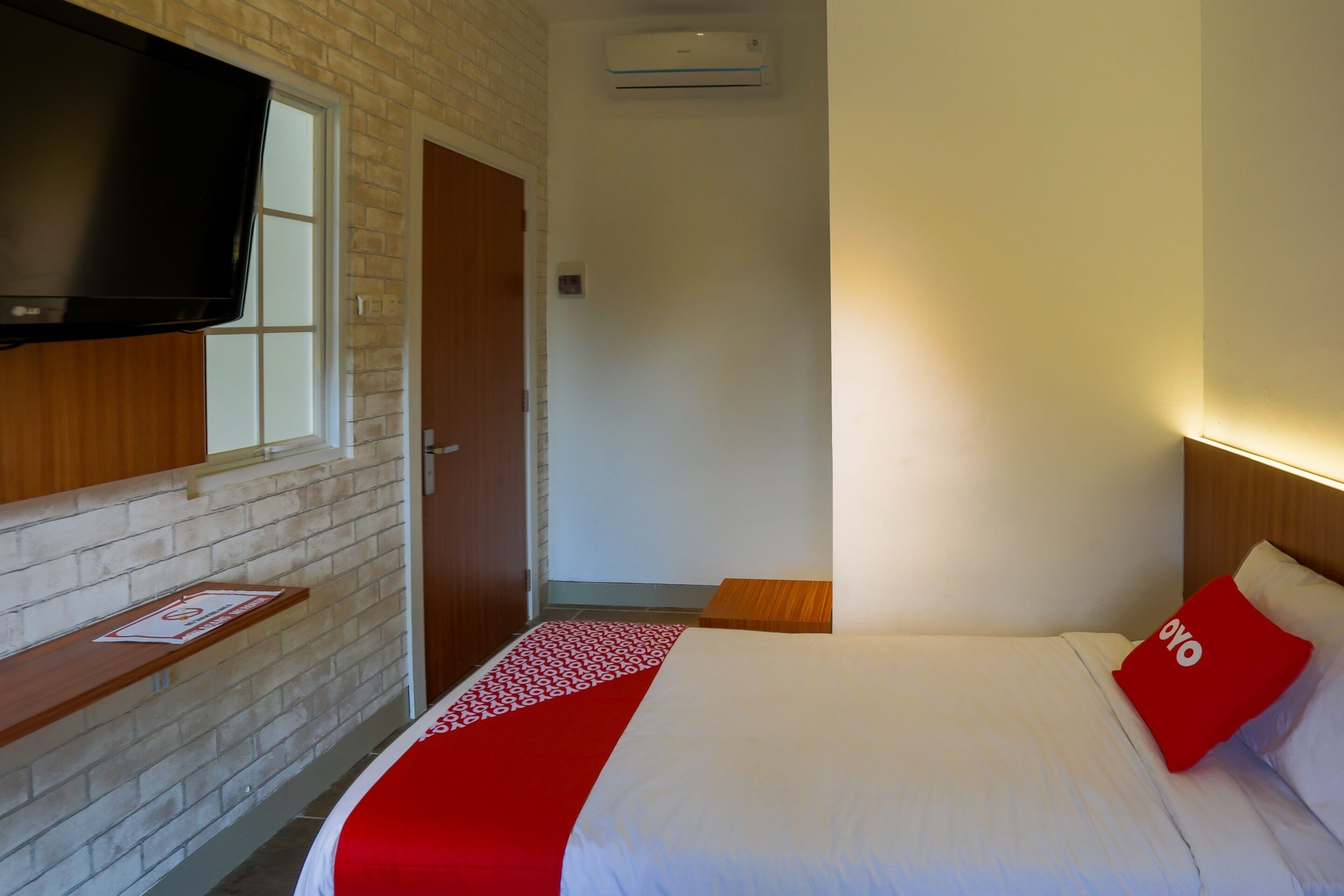 Ein Hotelzimmer mit Backsteinwand, einem flachbildfernseher über einem Holzregal, einem Bett mit weißer Bettwäsche und einem roten Kissen sowie einem kleinen Holzschemel.