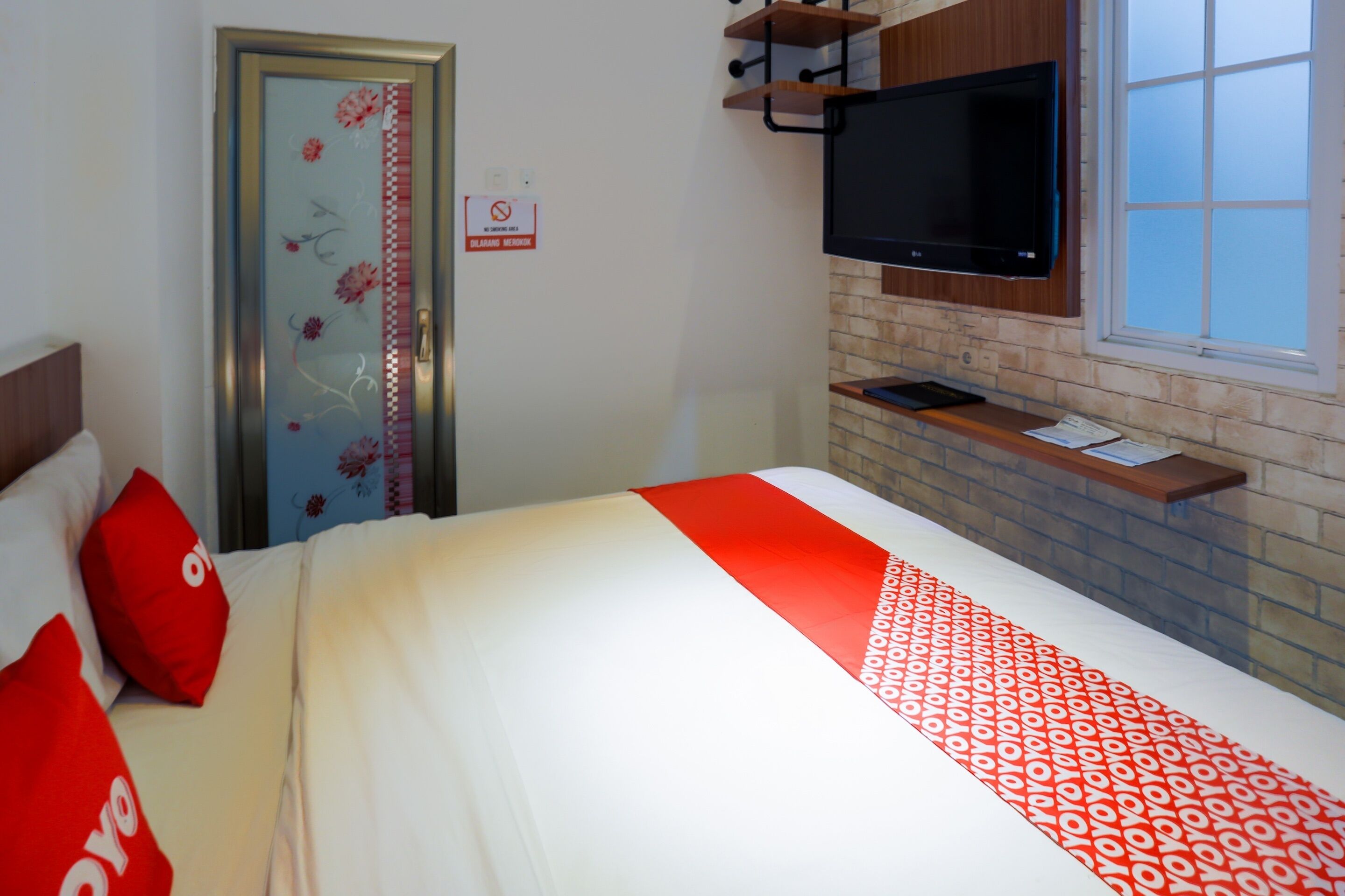 Une chambre d’hôtel avec un lit, un téléviseur fixé au mur, une fenêtre et une porte ornée de motifs floraux.