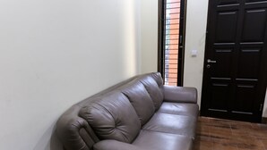 Ein braunes Ledersofa vor einer weißen Wand, im Hintergrund eine schwarze Tür und ein Fenster mit Metallgitter.