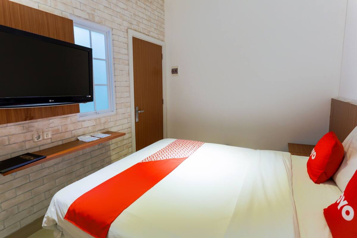 A hotel room with a flat-screen TV mounted on a brick wall, a bed with white bedding and red accents, and a wooden nightstand.
