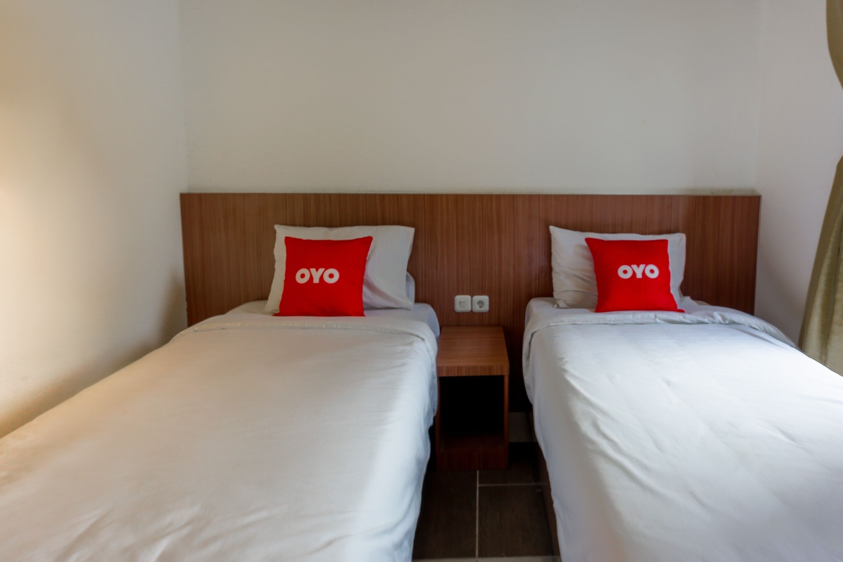 A hotel room with two single beds, each with a white mattress and a red pillow with "OYO" branding.