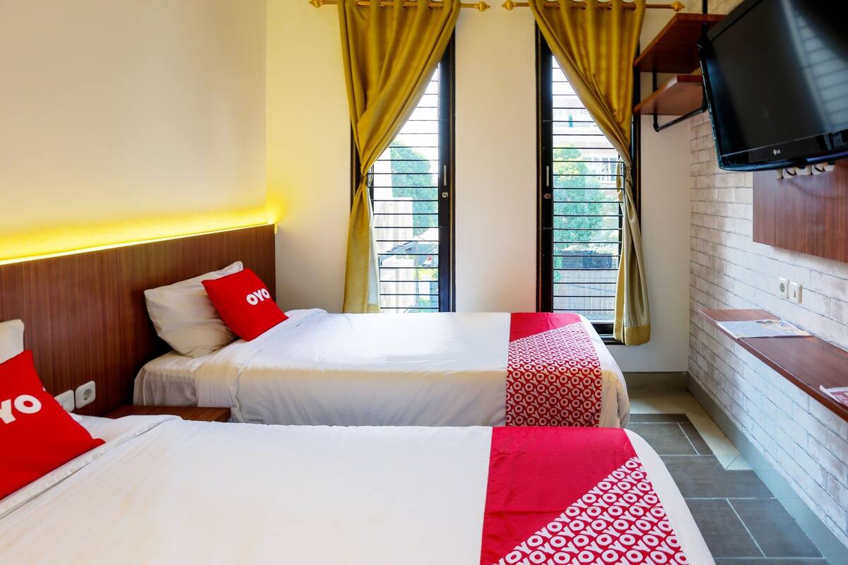 A hotel room with two beds, each with red and white bedding and pillows, a wooden headboard, a TV mounted on the wall, and a window with yellow curtains.