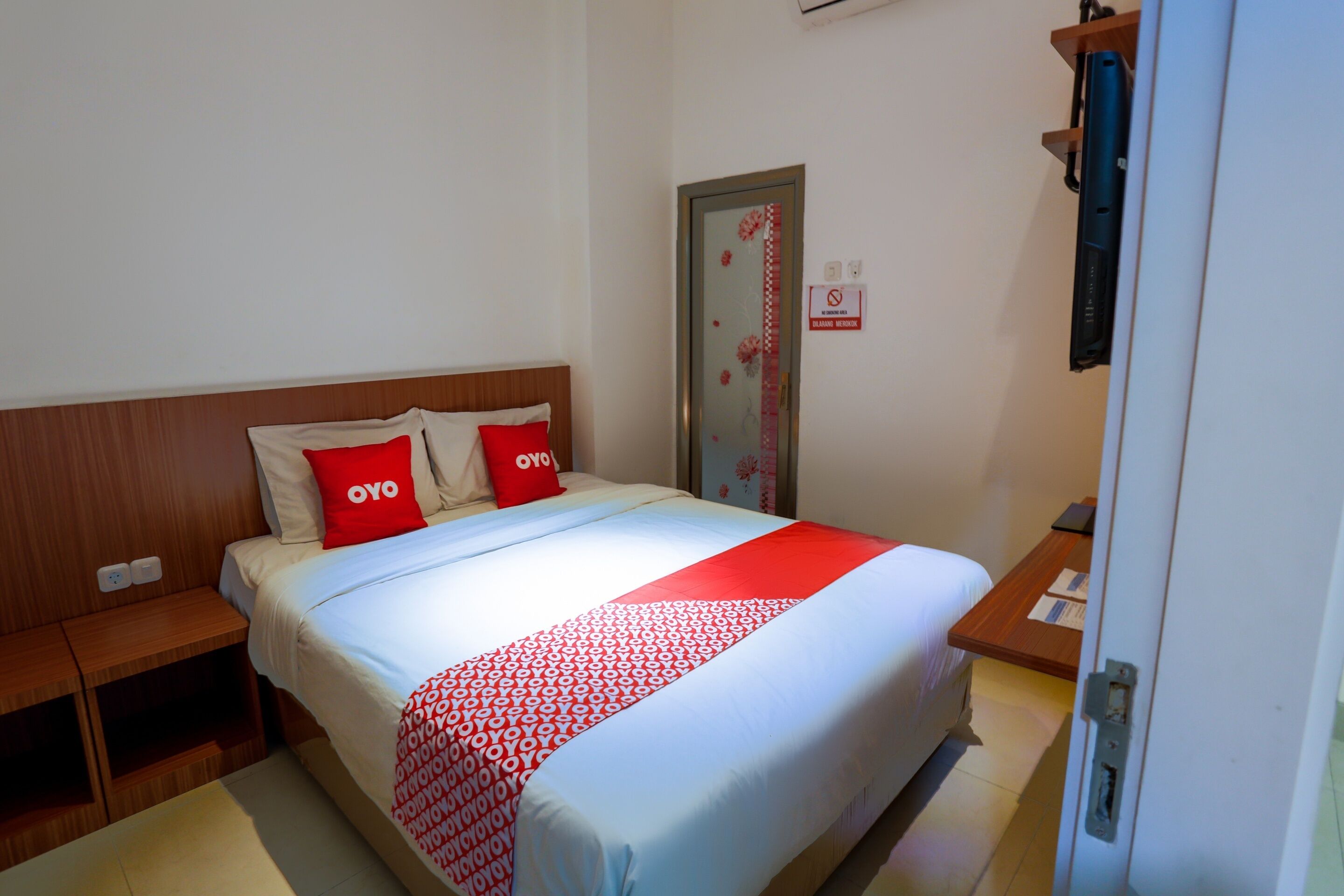 Habitación de hotel con cama, dos almohadas, manta roja, cabecera de madera, mesita de noche, puerta con cartel rojo y blanco, y una pequeña mesa con un libro y un bolígrafo.