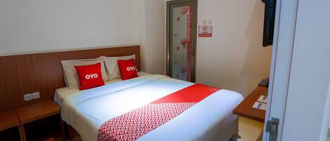 Habitación de hotel con cama, dos almohadas, manta roja, cabecera de madera, mesita de noche, puerta con cartel rojo y blanco, y una pequeña mesa con un libro y un bolígrafo.