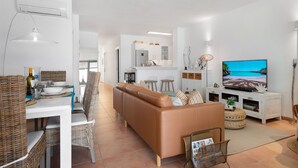 A modern living room with a brown leather sofa, a dining table with wicker chairs, a kitchen in the background, and a TV displaying a beach scene.
