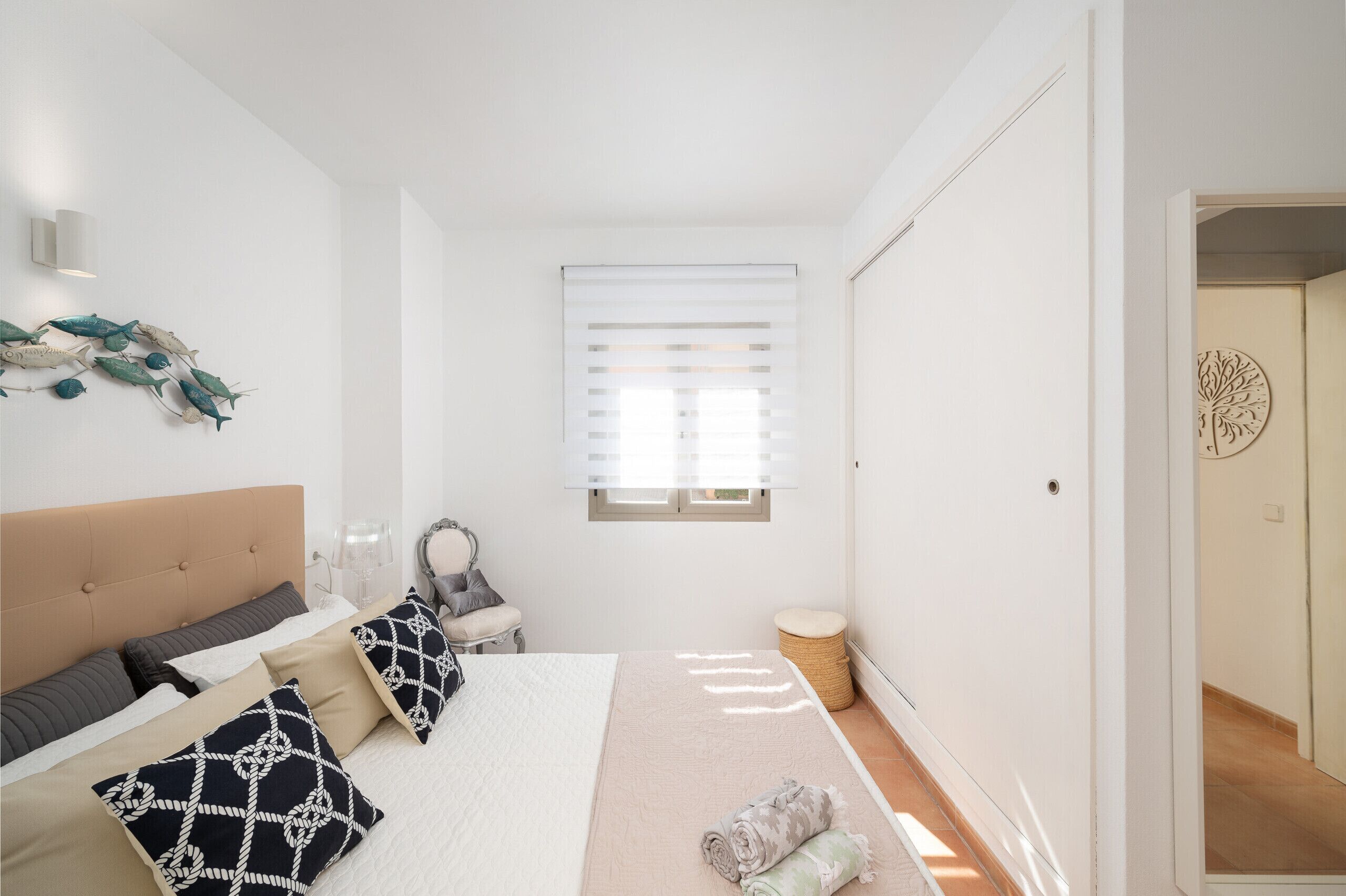 A bedroom with a bed, pillows, a bedside table, a lamp, a window with blinds, and a wall decoration.