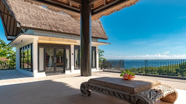 A modern building with a thatched roof, a balcony with a view of the sea, and a stone bench with a flower vase. - Villa Jempiring (Banjar)
