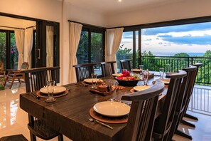 A dining area with a large table set for four, surrounded by chairs, with a view of greenery and a balcony. - Villa Jempiring (Banjar)