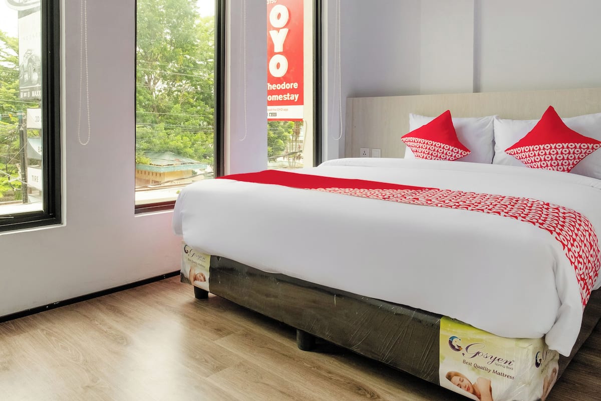 A hotel room with a large bed, red and white bedding, a view through the window, and a sign for 'OYO Homestay'.