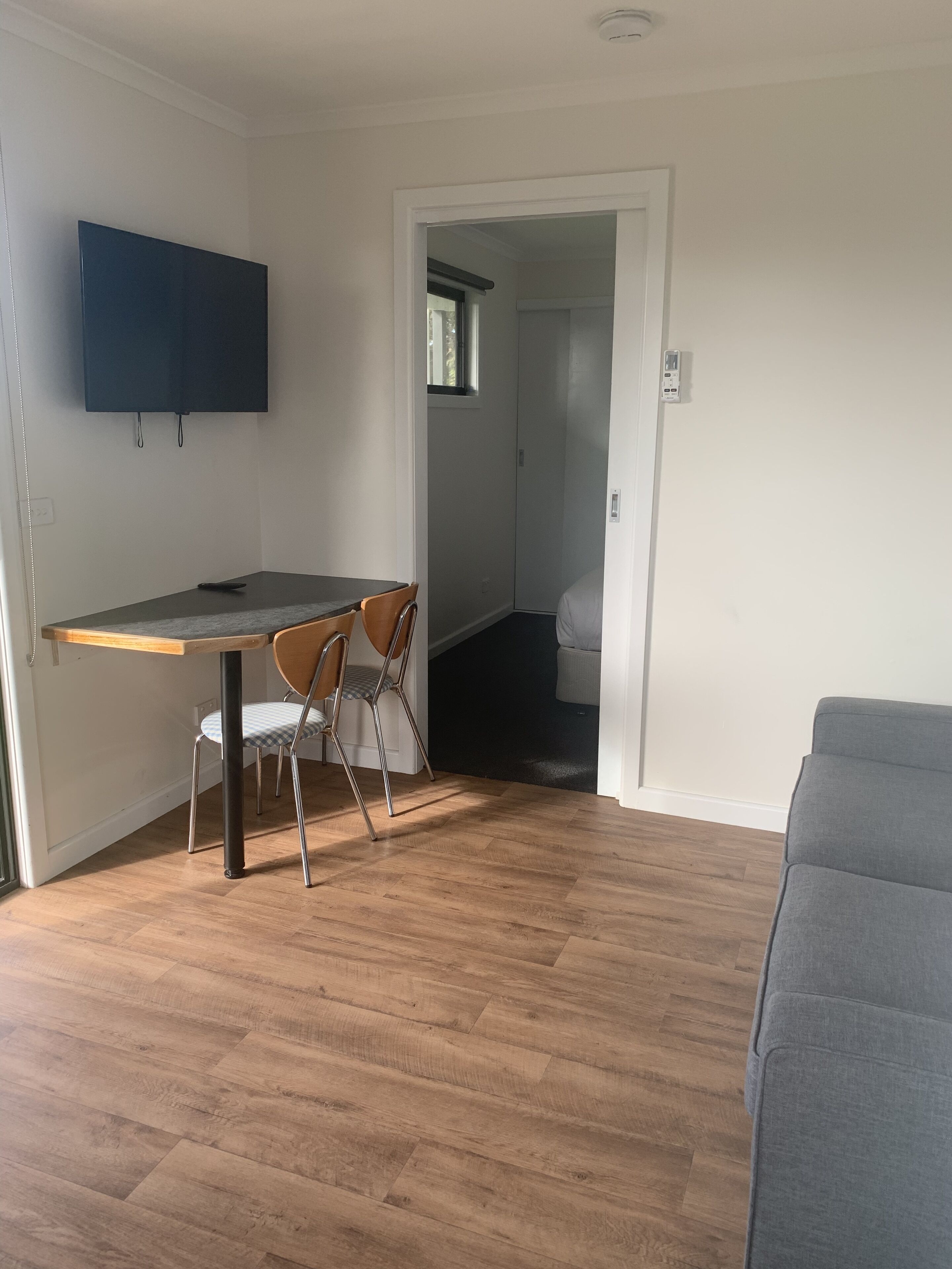 A room with a wooden dining table, two chairs, a grey sofa, and a TV mounted on the wall.