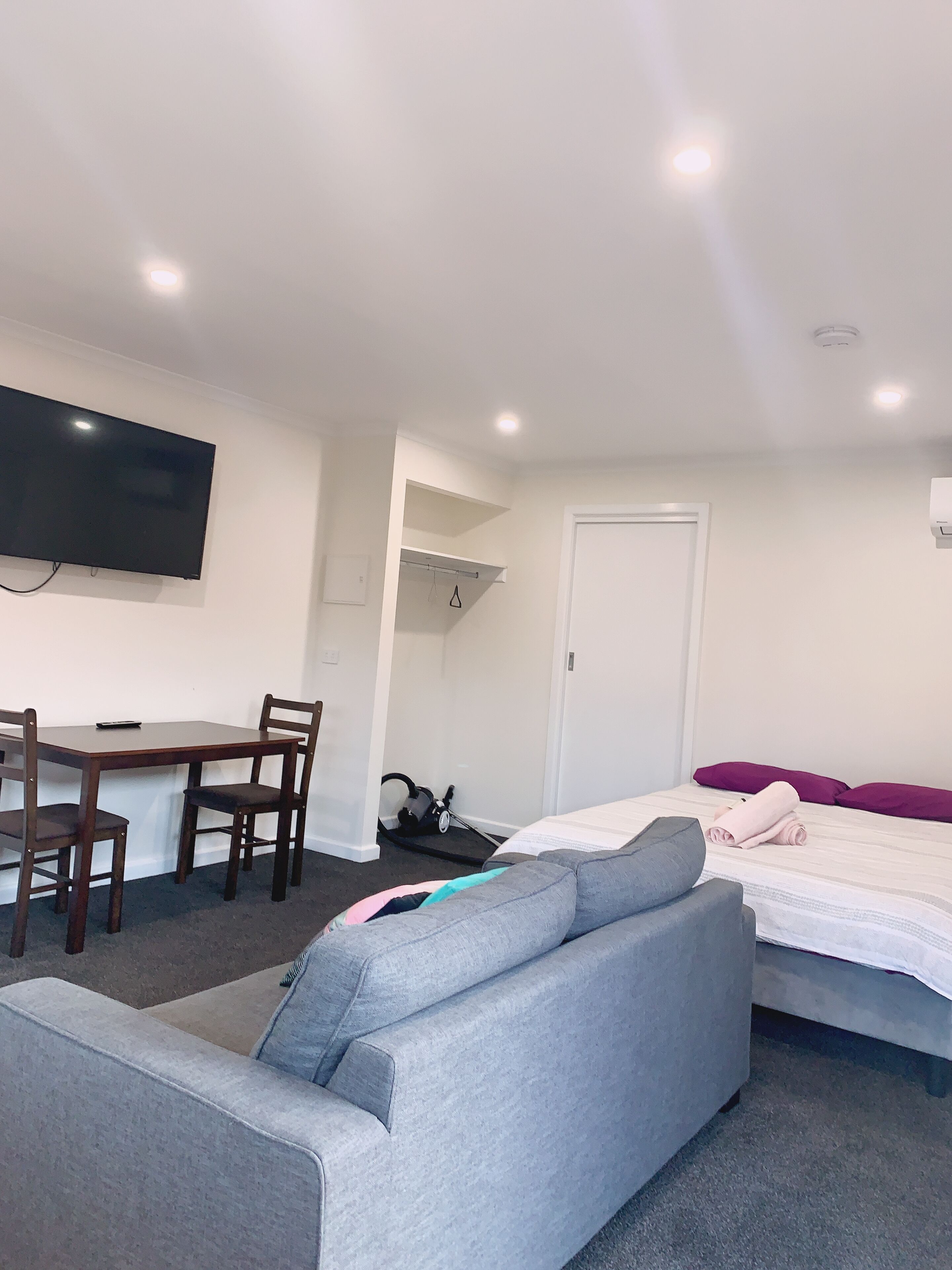 Et moderne hotelværelse med en seng, en grå sofa, et spisebord med stole, et fjernsyn ophængt på væggen og en dør.