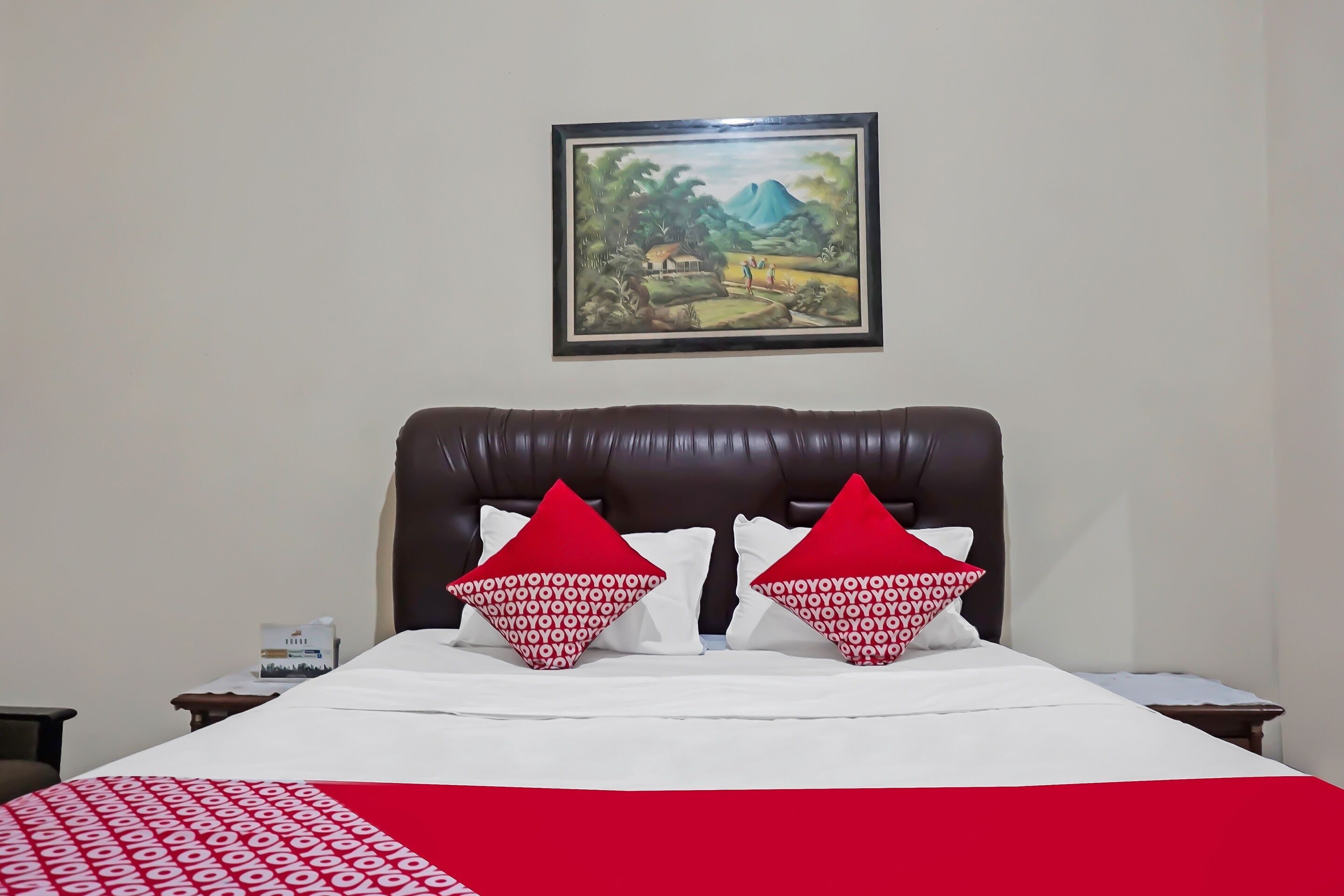 A hotel room with a large bed, two red patterned pillows, a white bedspread, and a framed landscape painting on the wall.