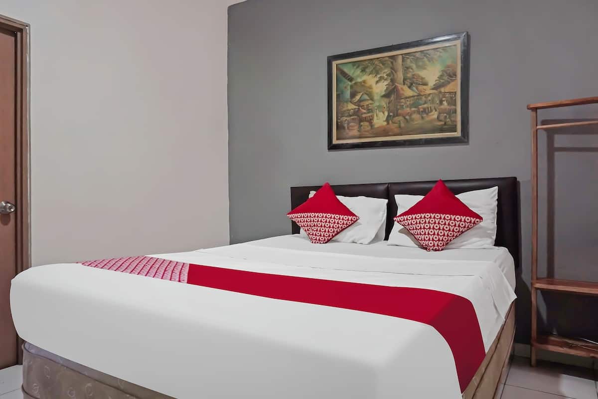 Een net opgemaakt bed met een wit-rode sprei, twee rode kussens en een houten hoofdbord. Daarboven hangt een schilderij in een lijst aan de muur.