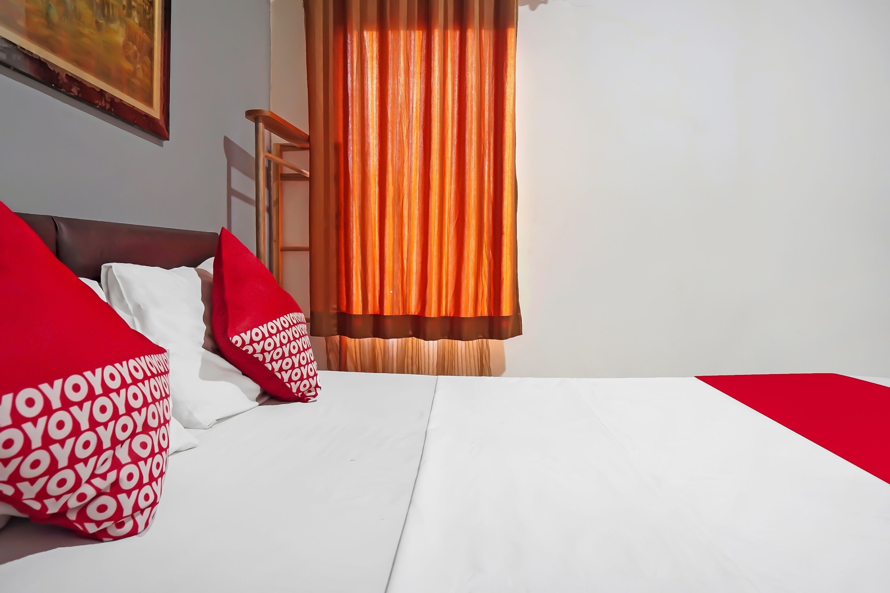 A hotel room with a bed, red and white pillows, a wooden headboard, a window with orange curtains, and a painting on the wall.
