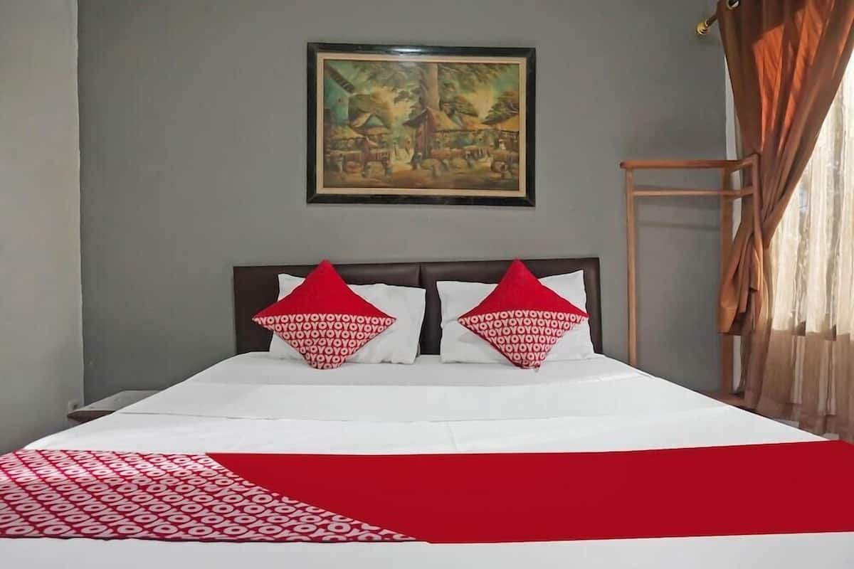 Een bed met een deken met een wit en rood patroon, twee rode kussens met een herhalend ontwerp, en een schilderij in een lijst aan de muur.