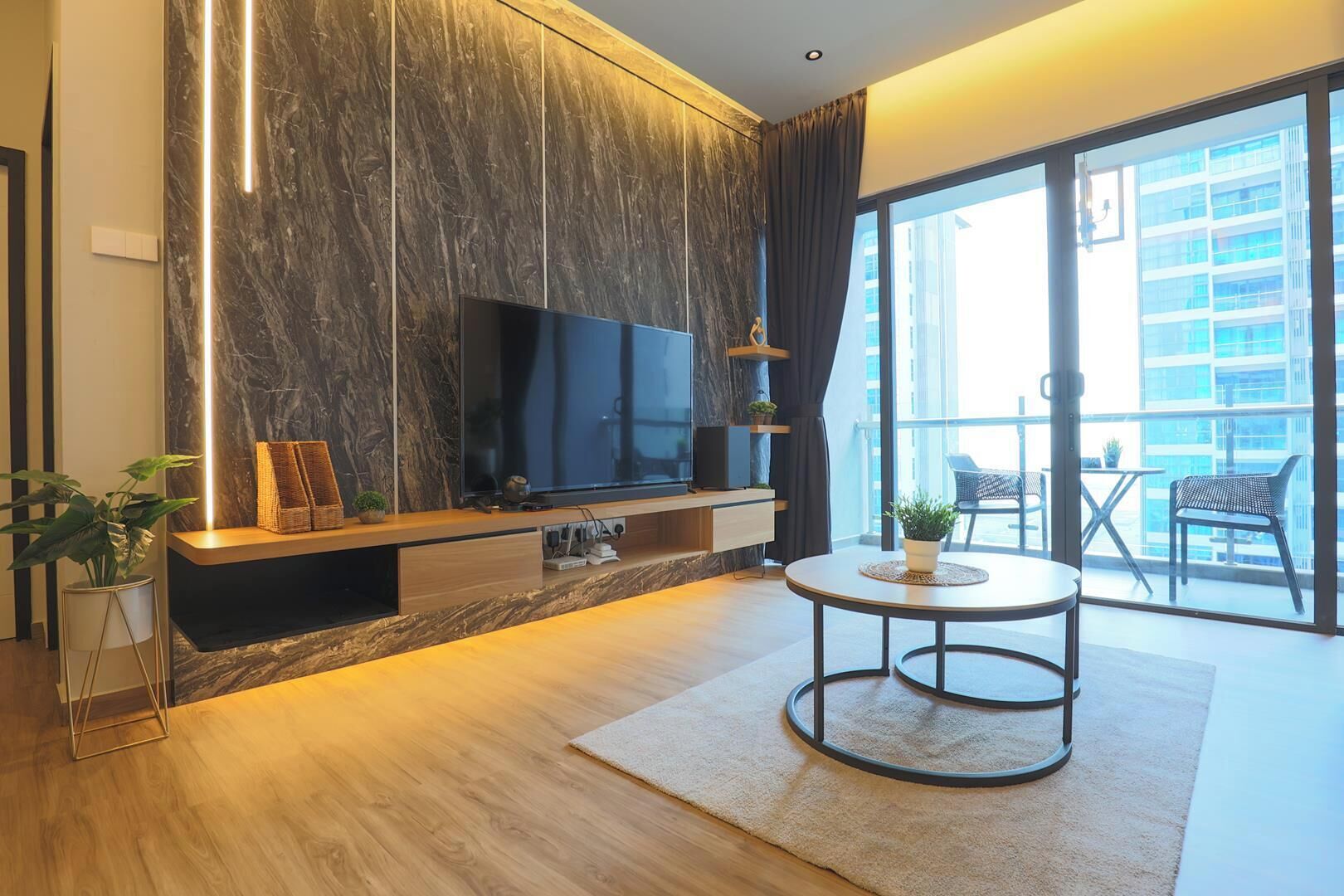 A modern living room with a flat-screen TV, wooden flooring, a round coffee table, and a balcony with outdoor furniture.