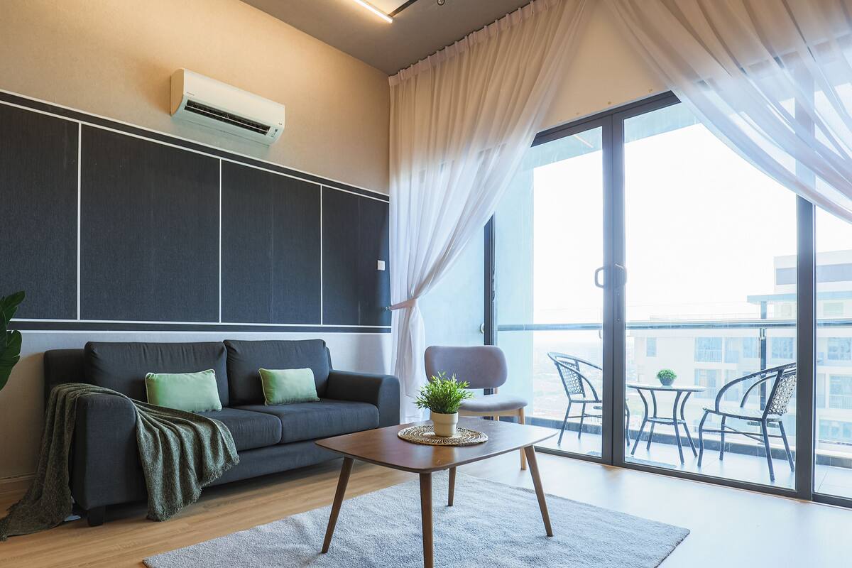Een moderne woonkamer met een donkergrijze bank, een klein houten bijzettafeltje en een groot raam met dunne gordijnen.
