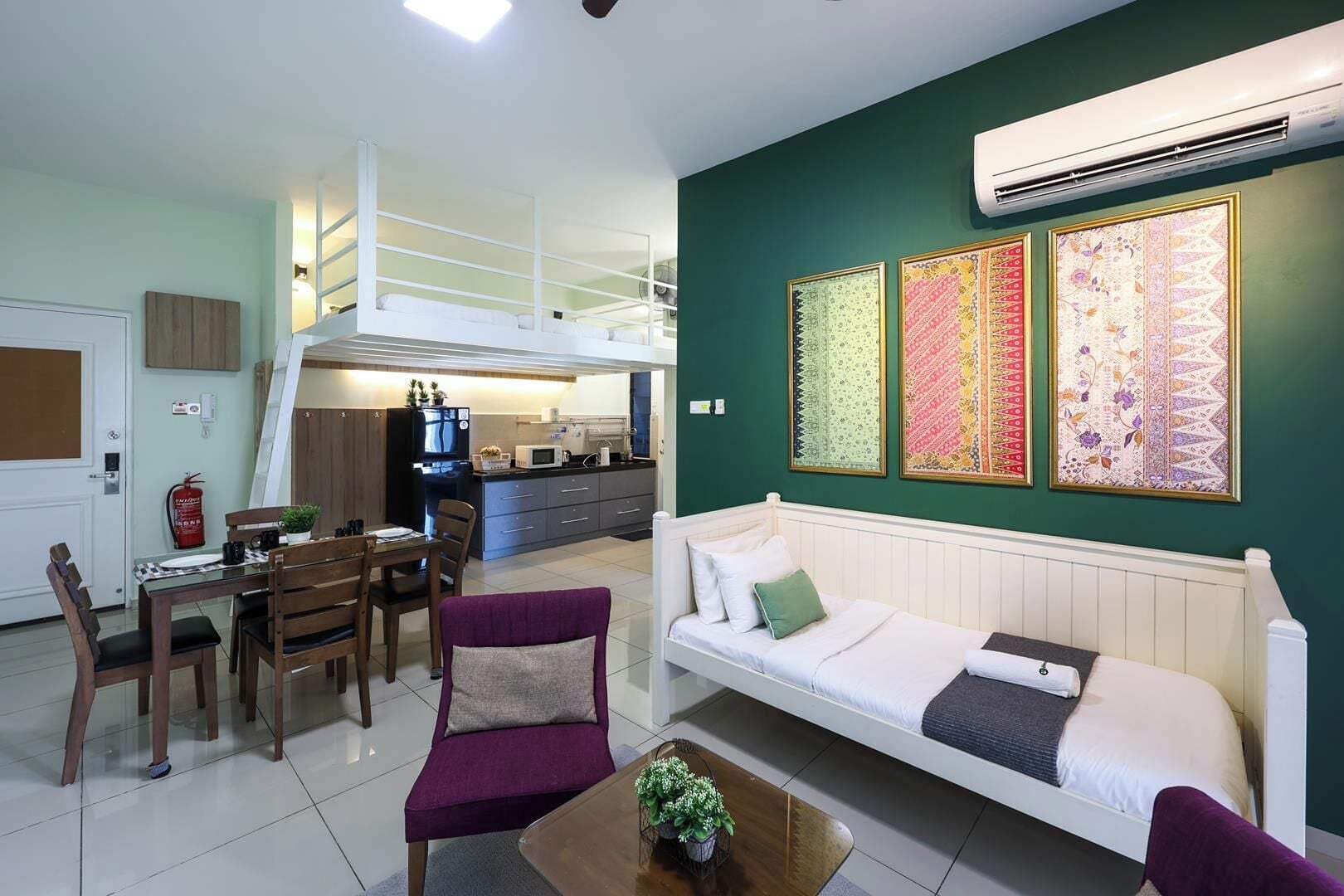 A modern living room with a sofa, a dining table, and chairs. There is a kitchen area with a refrigerator and microwave. The walls are painted green, and there are three framed artworks above the sofa.