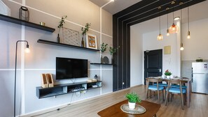 Una sala moderna con una televisión de pantalla plana, estantes con objetos decorativos, un comedor con sillas azules y una cocina con refrigerador.