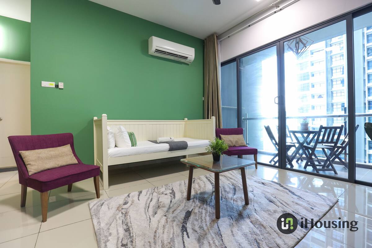Een moderne woonkamer met een groene wand, een witte bank, paarse fauteuils en een glazen salontafel.