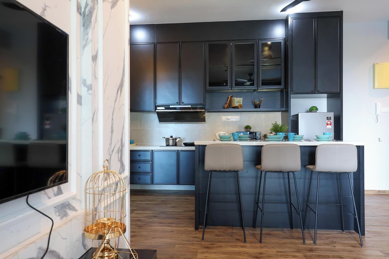 A modern kitchen with dark cabinets, a central island with bar stools, and a flat-screen TV mounted on the wall.