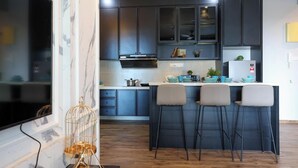 A modern kitchen with dark cabinets, a central island with bar stools, and a flat-screen TV mounted on the wall. - Atlantis Residence Premium By I Housing (Malacca City)