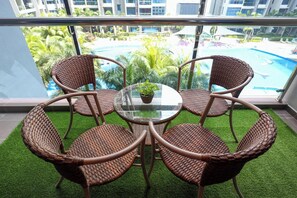 A balcony with wicker chairs and a glass table, overlooking a swimming pool and greenery.