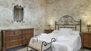Une chambre avec un mur en pierre, un lit avec du linge de lit blanc, une commode en bois, une chaise et un miroir.